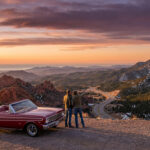 Classic red convertible and two travelers overlook a vast American landscape with red rocks, snowy mountains, and ocean at golden hour.