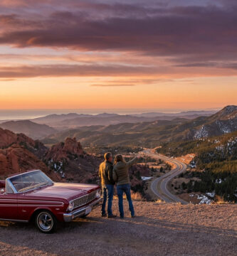 Classic red convertible and two travelers overlook a vast American landscape with red rocks, snowy mountains, and ocean at golden hour.
