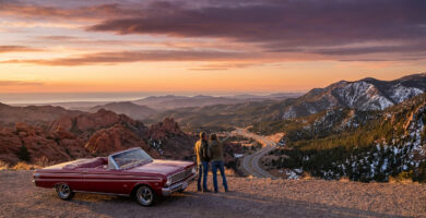 Classic red convertible and two travelers overlook a vast American landscape with red rocks, snowy mountains, and ocean at golden hour.