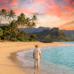 Personne en chapeau sur une plage de sable fin à Hawaï, face à un coucher de soleil rose-orange, palmiers et montagnes.