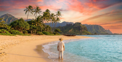 Personne en chapeau sur une plage de sable fin à Hawaï, face à un coucher de soleil rose-orange, palmiers et montagnes.