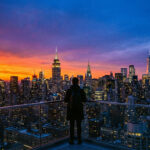 Vue aérienne de New York au crépuscule. Un homme de dos sur un balcon admire la skyline illuminée avec un ciel orange et bleu intense.