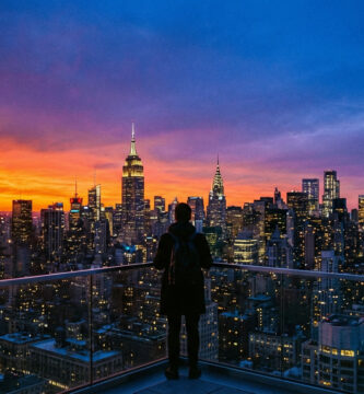 Vue aérienne de New York au crépuscule. Un homme de dos sur un balcon admire la skyline illuminée avec un ciel orange et bleu intense.