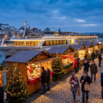 Un marché de Noël animé le long d'une rivière avec un bateau de croisière illuminé au crépuscule en arrière-plan.